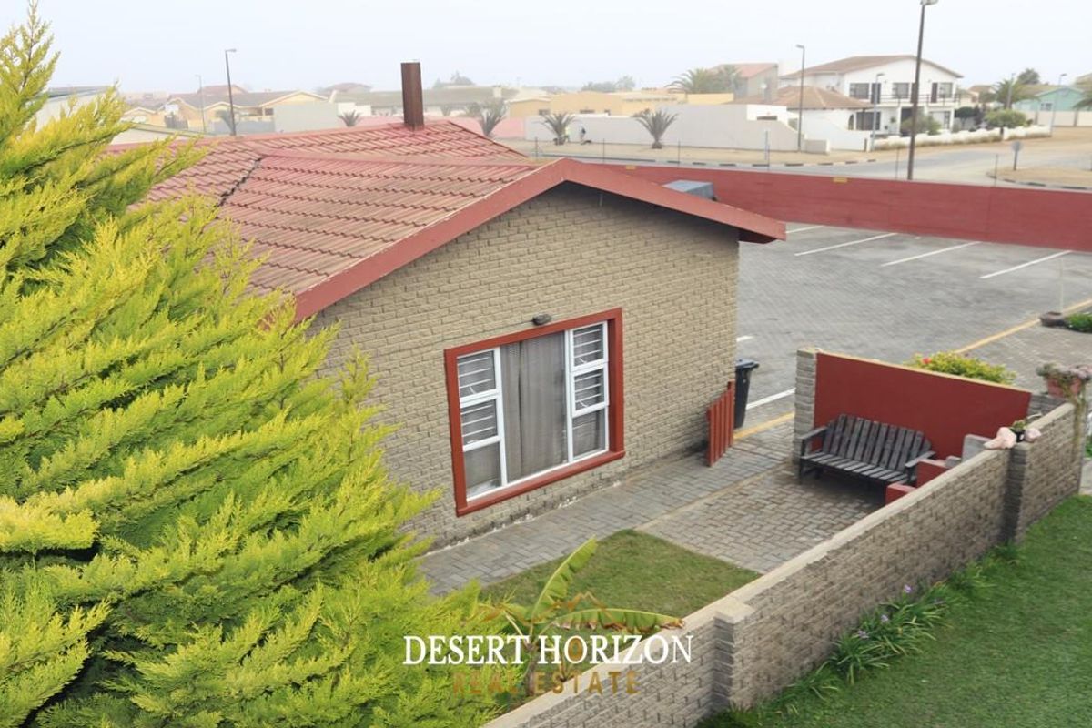 An elevated view of a single-story brick house featuring a tiled roof, paved patio area, and surrounding garden space within a residential complex.