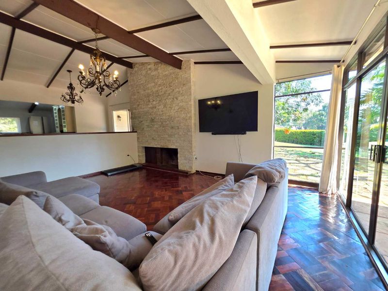 Spacious living room featuring exposed timber ceiling beams, a prominent stone-clad fireplace, and classic parquet flooring with large glass doors opening to the garden.