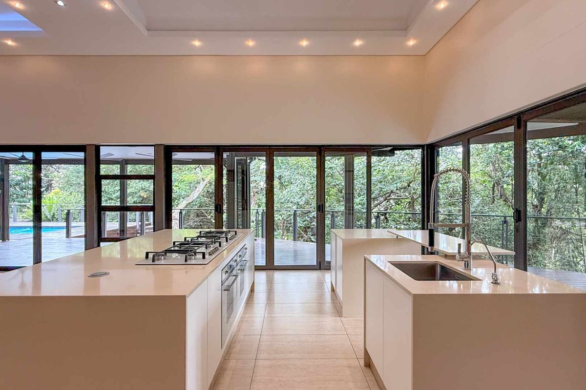Modern open-plan kitchen featuring dual islands, stone countertops, integrated gas hob, and expansive floor-to-ceiling glass doors opening to a lush outdoor deck.