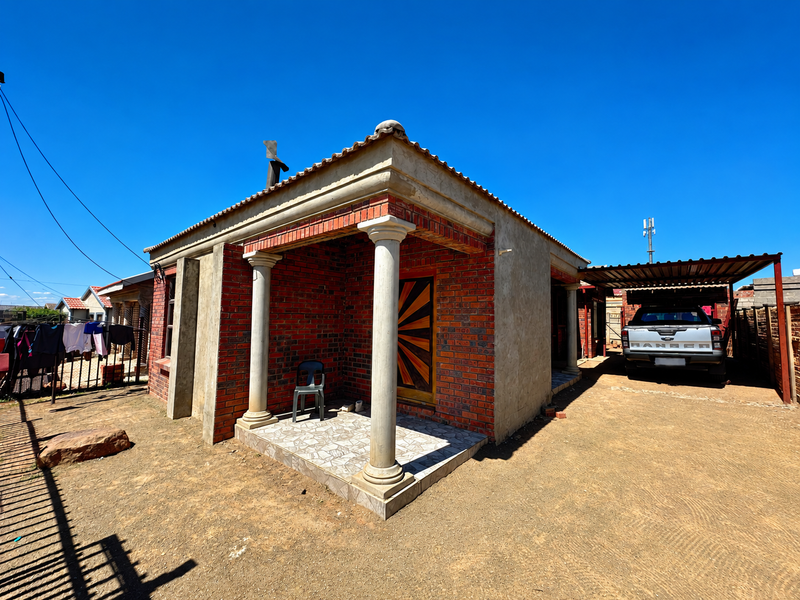 Front view of a single-story brick house featuring a covered porch with concrete columns, a side carport, and an unfinished exterior wall finish.