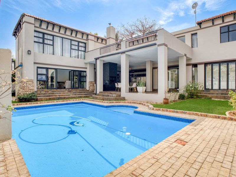 Spacious rear exterior featuring a swimming pool, paved patio area, covered entertainment space with built-in fireplace, and a balcony overlooking the garden.
