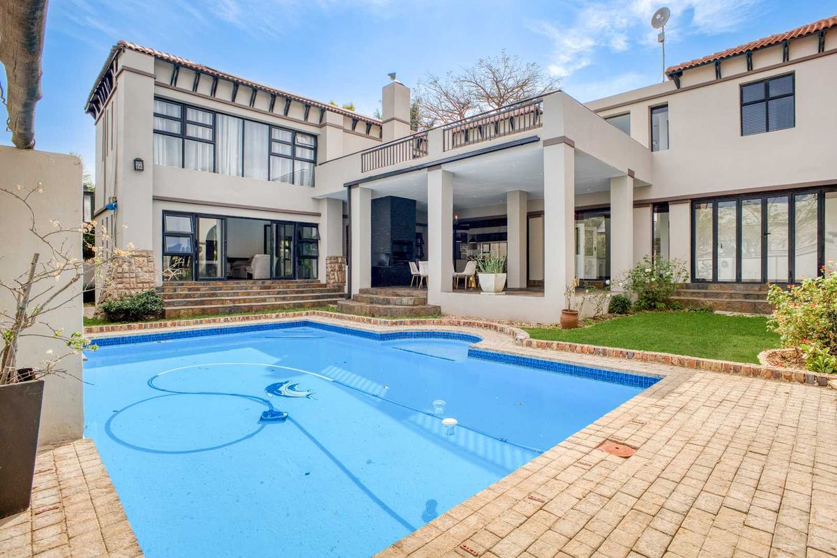 Spacious rear exterior featuring a swimming pool, paved patio area, covered entertainment space with built-in fireplace, and a balcony overlooking the garden.