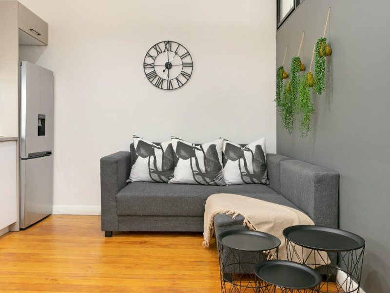 A modern open-plan living area featuring hardwood flooring, a compact kitchen with white cabinetry, and a neutral grey accent wall.