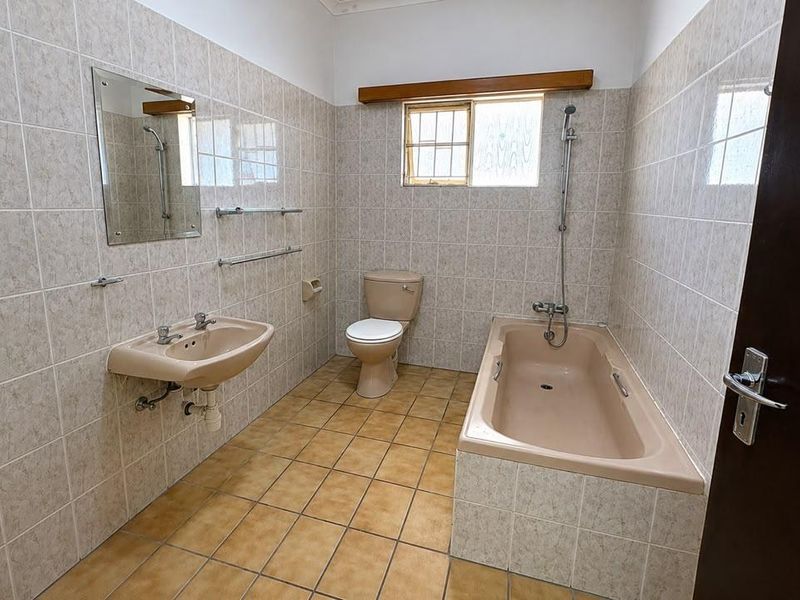 A dated bathroom featuring a built-in bathtub, wall-mounted basin, and toilet, finished with neutral wall tiles and terracotta-style floor tiles.