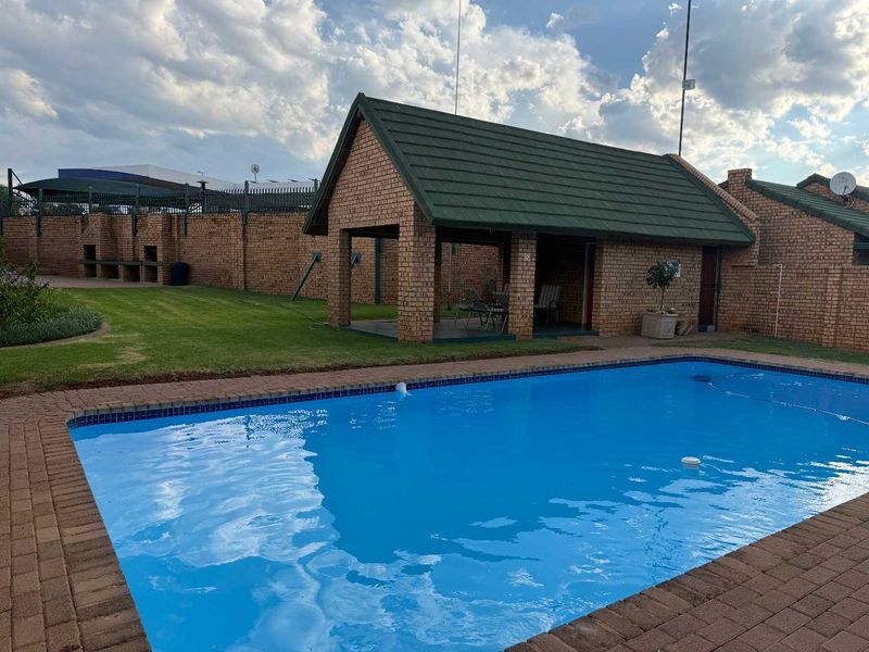 A well-maintained outdoor pool area featuring a paved surround, a covered patio structure, and a built-in braai area within a secure brick-walled garden.