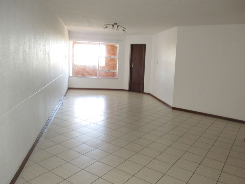 A spacious, empty living area featuring neutral tiled flooring, a ceiling-mounted light fixture, and a large window providing natural light.