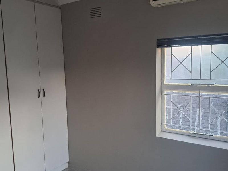 A functional bedroom featuring built-in cupboards, tiled flooring, and a wall-mounted air conditioning unit with security bars on the window.