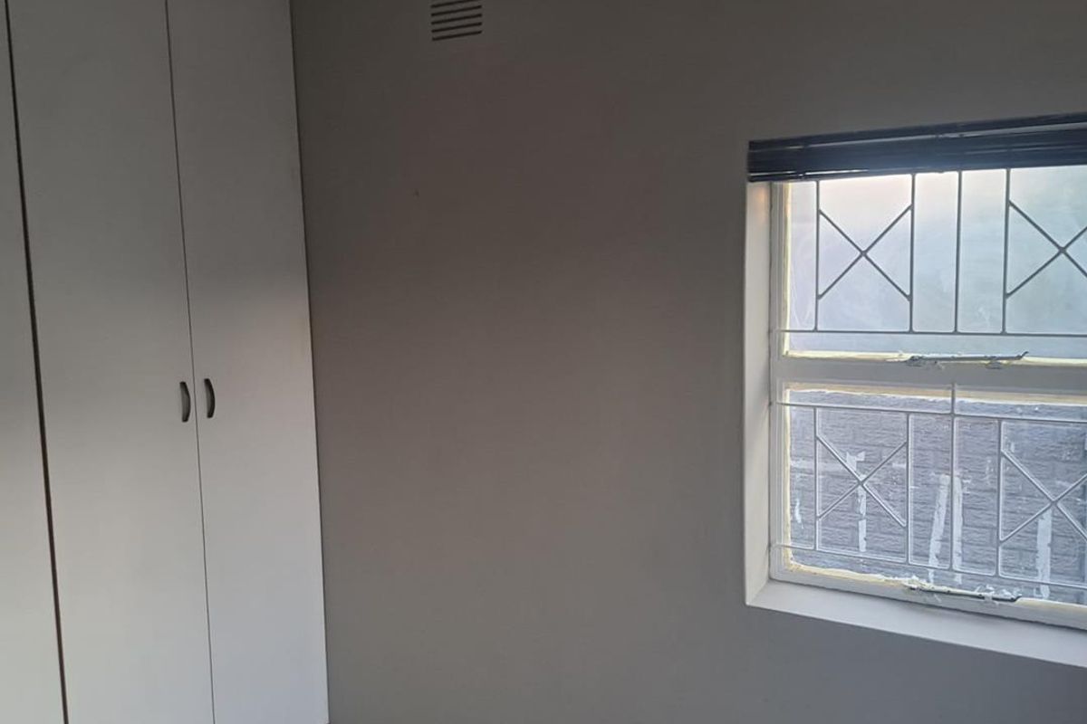 A functional bedroom featuring built-in cupboards, tiled flooring, and a wall-mounted air conditioning unit with security bars on the window.