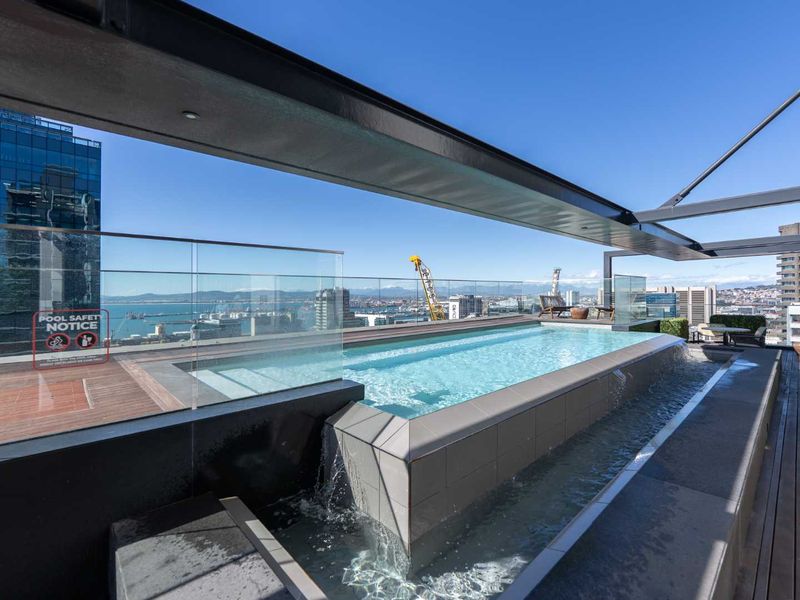 A modern rooftop infinity pool featuring glass balustrades, timber decking, and a steel pergola, offering expansive city and harbour views.