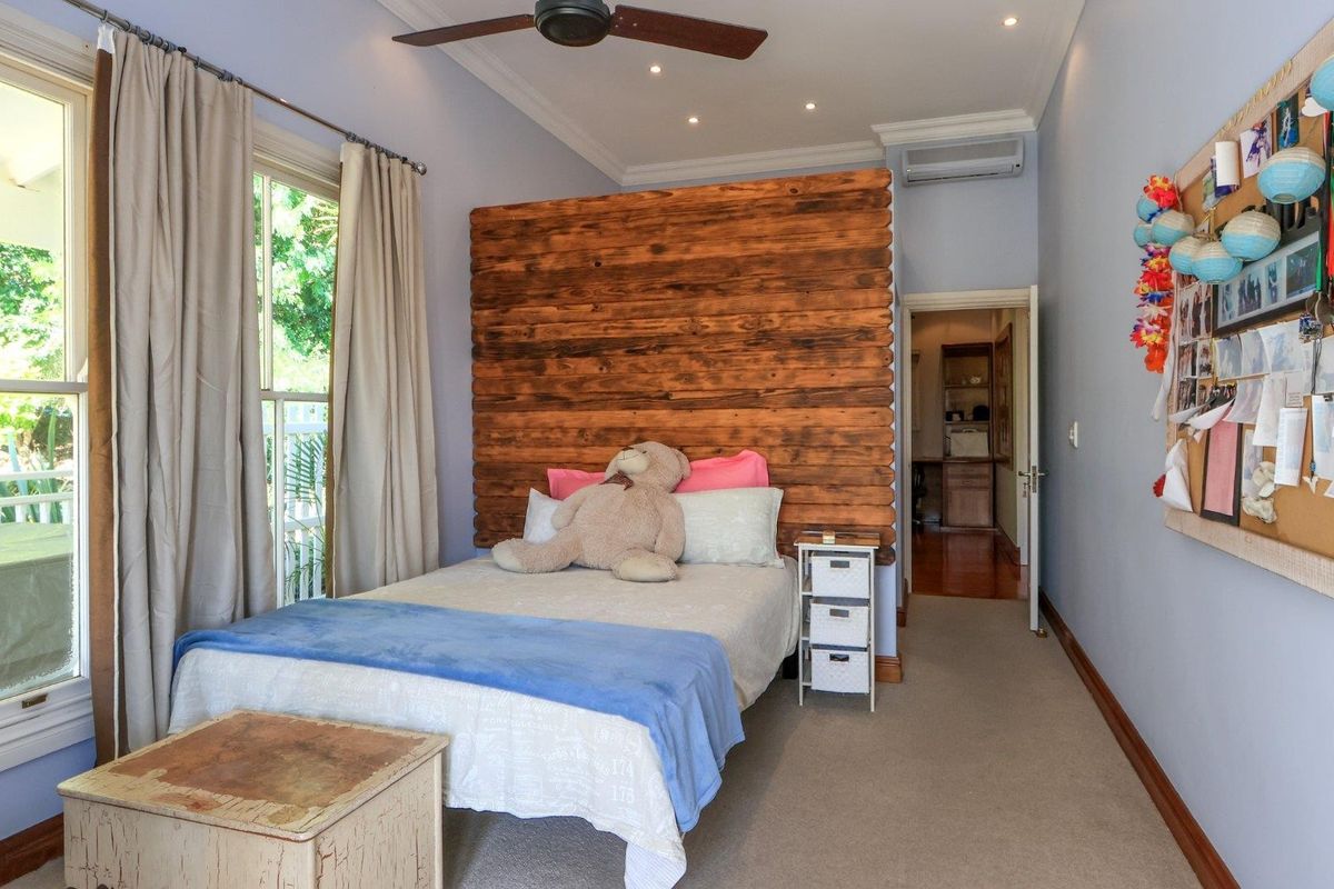 A well-maintained bedroom featuring a timber accent wall, ceiling fan, split-system air conditioning, and neutral carpet flooring with access to an adjoining room.