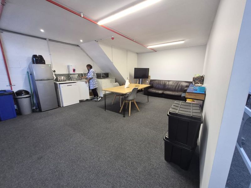 An open-plan office kitchenette area featuring carpeted flooring, exposed fire sprinkler piping, and functional cabinetry with a tiled backsplash.