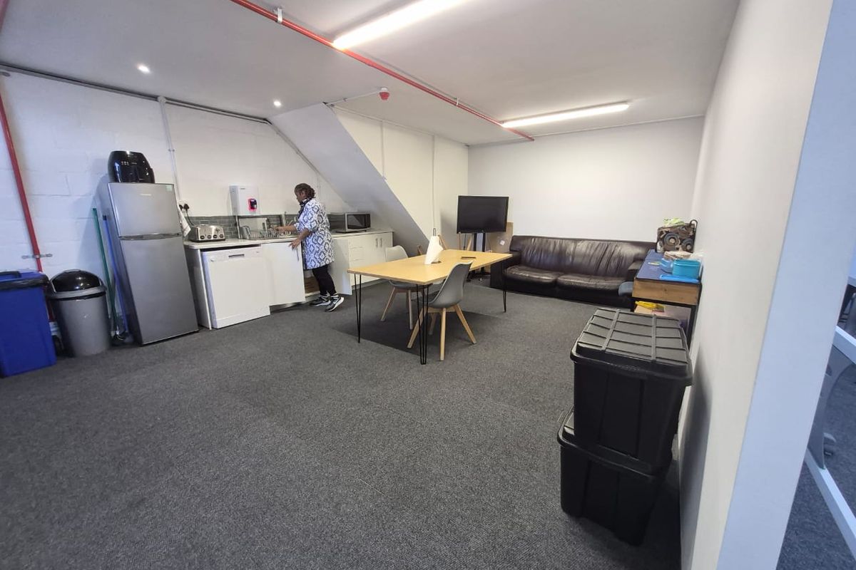 An open-plan office kitchenette area featuring carpeted flooring, exposed fire sprinkler piping, and functional cabinetry with a tiled backsplash.