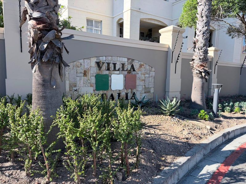 Front entrance view of the Villa Italia apartment complex, featuring a stone-clad signage wall, established palm trees, and secure perimeter fencing.