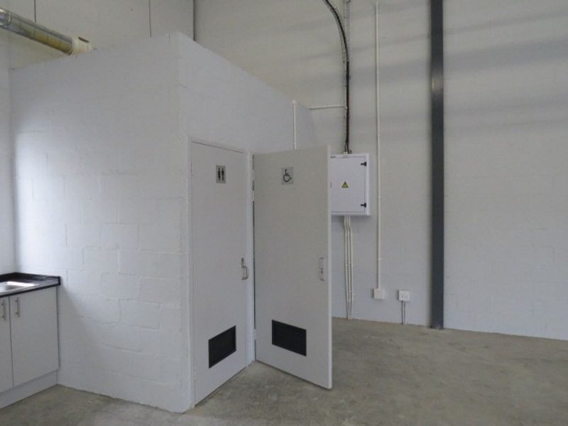 Industrial utility area featuring an accessible restroom, standard restroom, and wall-mounted electrical distribution board with exposed conduit and concrete flooring.
