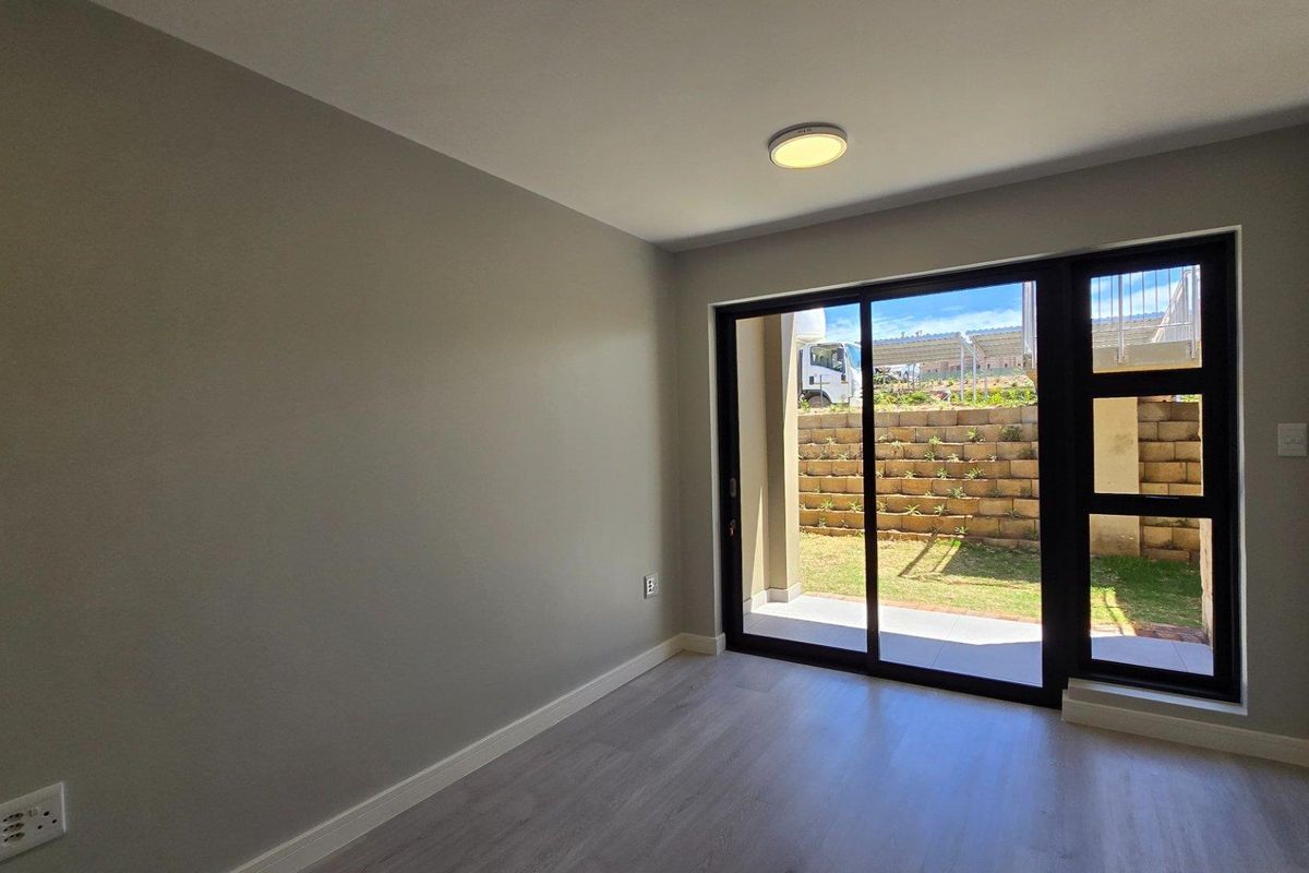A bright, empty bedroom featuring modern light-toned flooring, neutral walls, and large sliding glass doors that open onto a private patio and garden area.