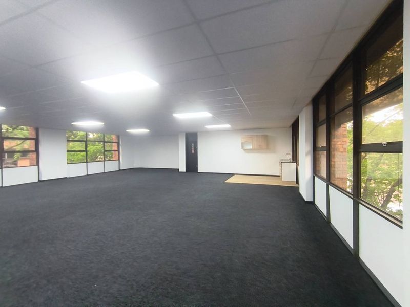 Spacious open-plan office area featuring large windows for natural light, recessed ceiling lighting, carpeted flooring, and a compact fitted kitchenette.