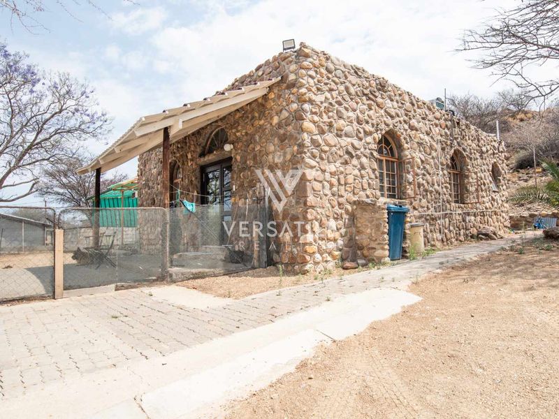 A rustic stone-clad cottage featuring arched windows and a covered patio area, set within a natural landscape on a small holding.