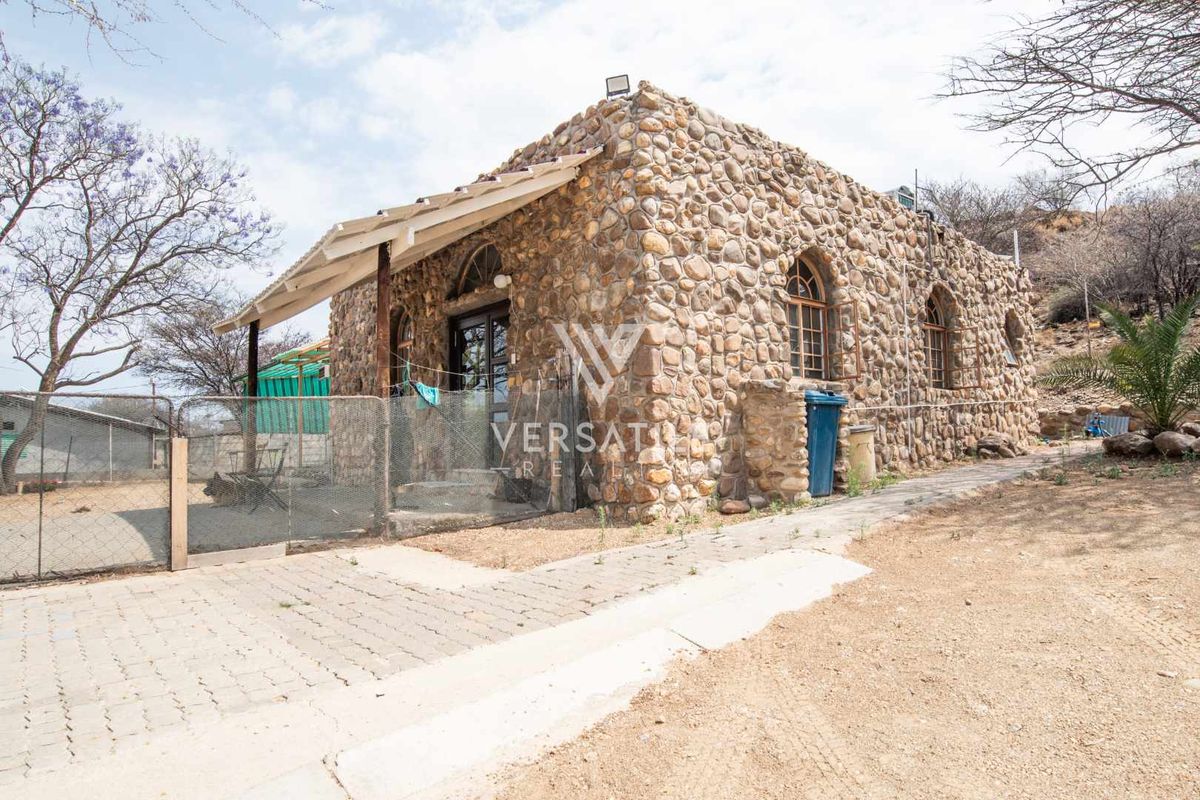 A rustic stone-clad cottage featuring arched windows and a covered patio area, set within a natural landscape on a small holding.