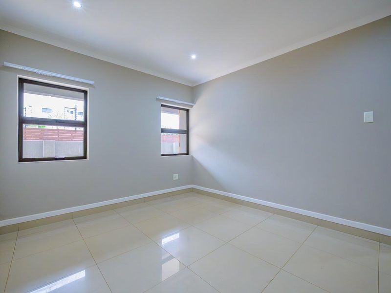 A bright, empty bedroom featuring neutral wall paint, polished tiled flooring, and modern aluminium-framed windows with recessed ceiling lighting.