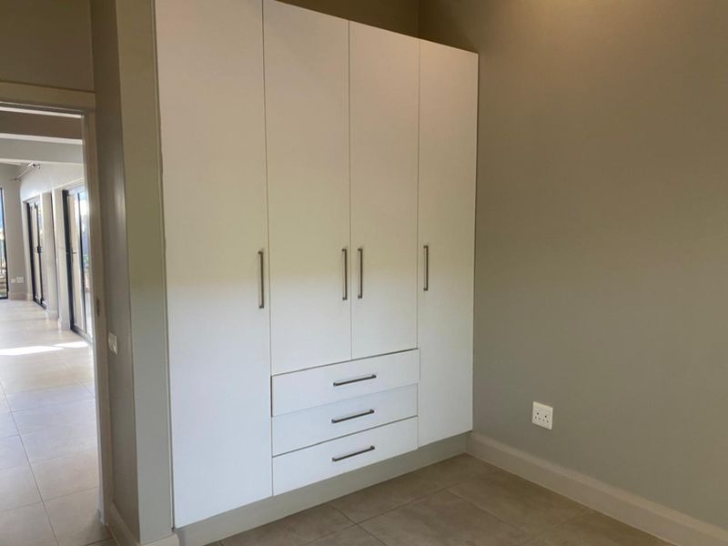 A clean, modern bedroom featuring neutral wall tones, tiled flooring, and a large, white built-in wardrobe with integrated drawers.