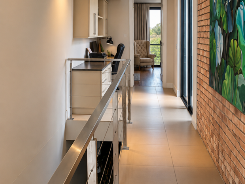 A modern hallway featuring a stainless steel balustrade, exposed brick feature wall, recessed lighting, and a built-in study nook with cabinetry.