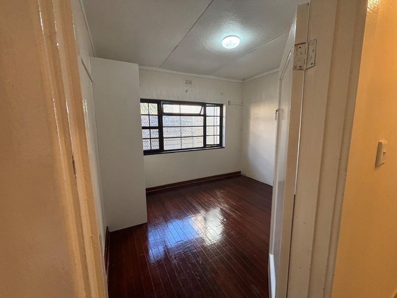 An empty bedroom featuring polished hardwood flooring, a built-in cupboard, and a window fitted with security bars.