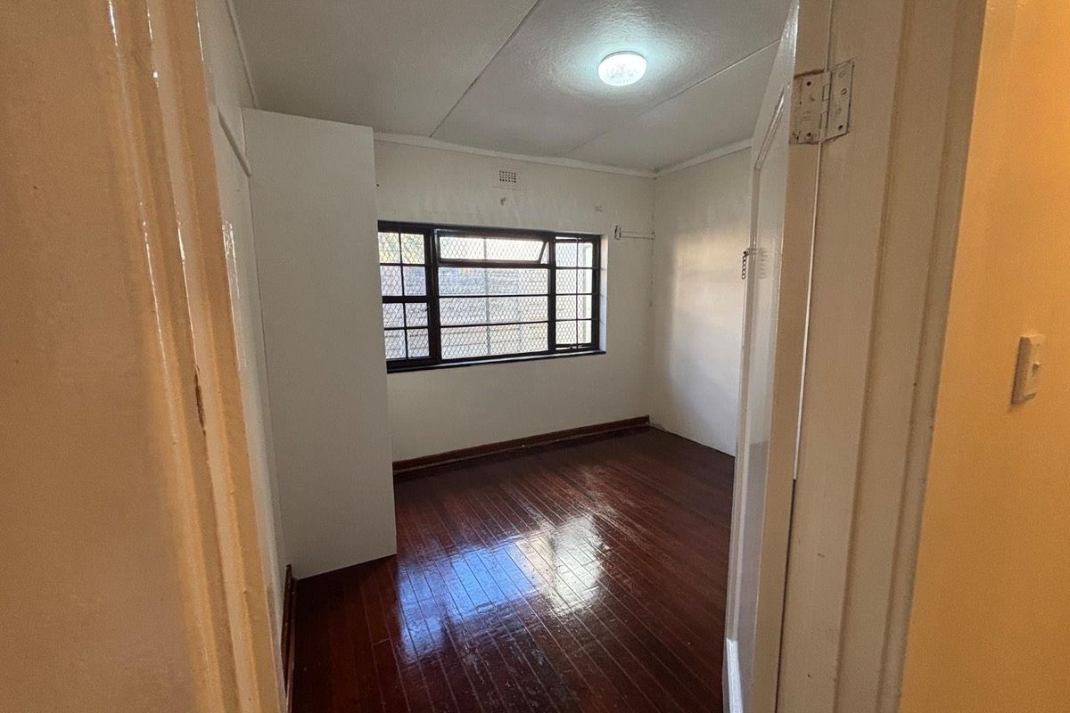 An empty bedroom featuring polished hardwood flooring, a built-in cupboard, and a window fitted with security bars.