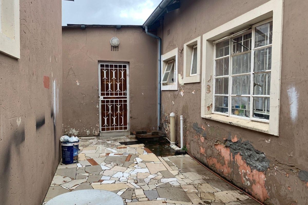 A rear exterior courtyard area featuring paved flooring, security gate, and windows, showing significant signs of wear and weathering on the exterior walls.