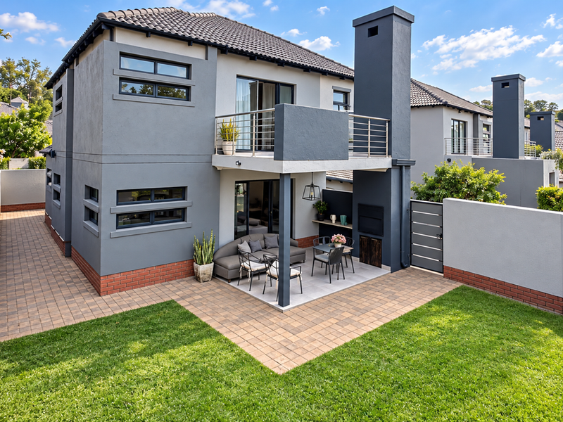Rear view of a modern townhouse featuring a paved patio, built-in braai, upper-level balcony with stainless steel balustrade, and a well-maintained private garden.