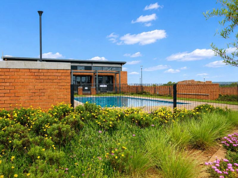 A well-maintained swimming pool area featuring a brick boundary wall, wire mesh fencing, and established garden landscaping.