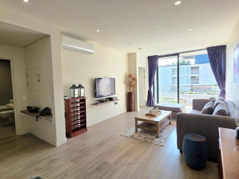 A bright, open-plan living area featuring laminate flooring, recessed lighting, and a split-system air conditioner, with direct access to a private balcony.