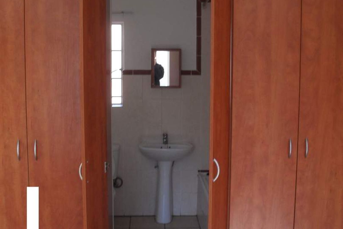 A view through built-in wooden cupboards into a small bathroom featuring a pedestal basin and tiled flooring with visible exposed plumbing.
