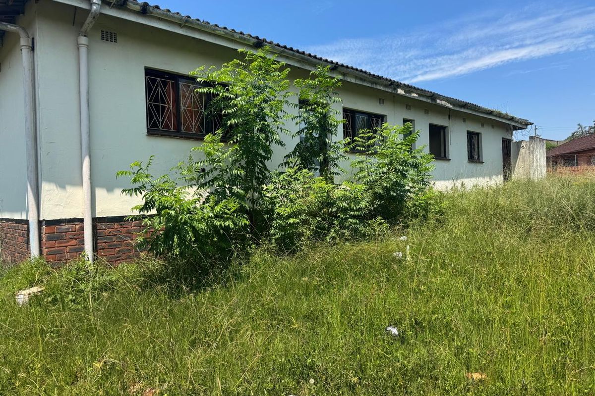 Rear exterior view of the property featuring a brick base, security bars on windows, and significant overgrown vegetation requiring maintenance.