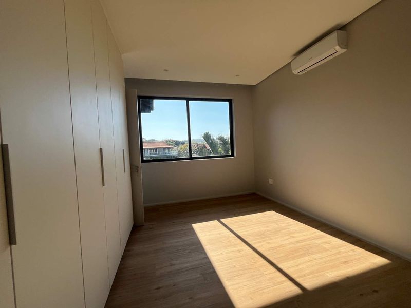 A bright, empty bedroom featuring modern laminate flooring, built-in cupboards, and a wall-mounted air conditioning unit with a pleasant exterior view.
