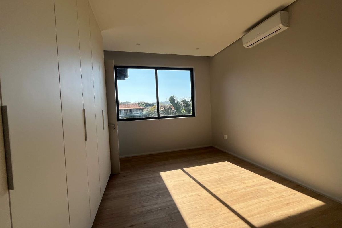 A bright, empty bedroom featuring modern laminate flooring, built-in cupboards, and a wall-mounted air conditioning unit with a pleasant exterior view.