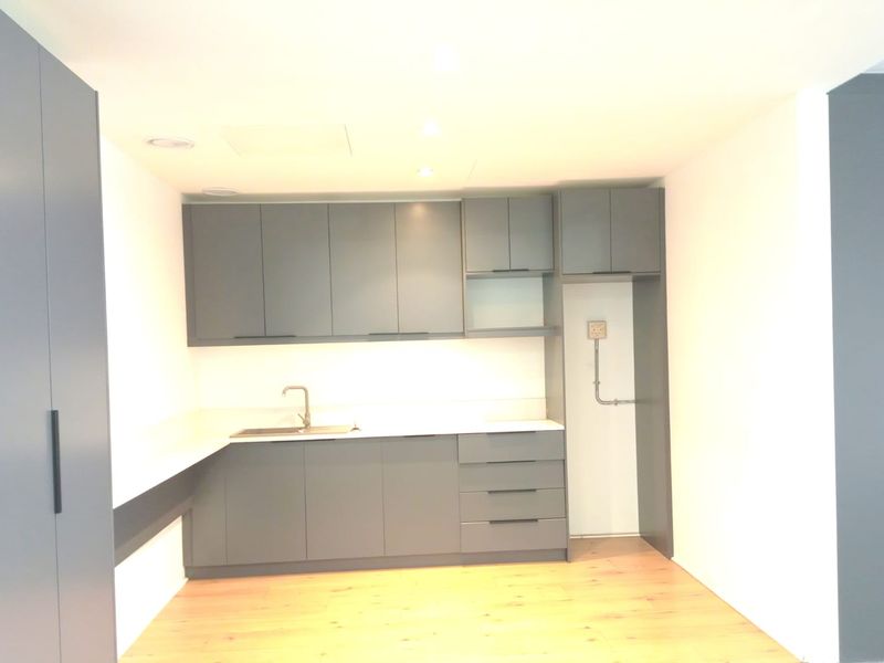A modern, minimalist kitchenette featuring sleek grey cabinetry, a stainless steel sink, and light wood-look flooring in a clean, bright office space.