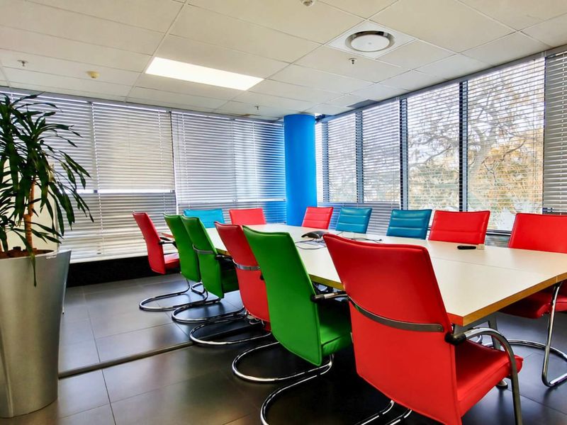 A bright office boardroom featuring large windows with venetian blinds, tiled flooring, and a suspended ceiling with integrated lighting.