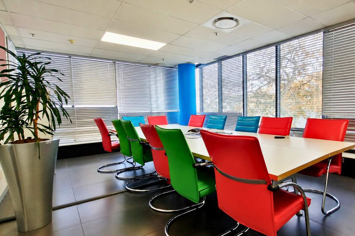 A bright office boardroom featuring large windows with venetian blinds, tiled flooring, and a suspended ceiling with integrated lighting.