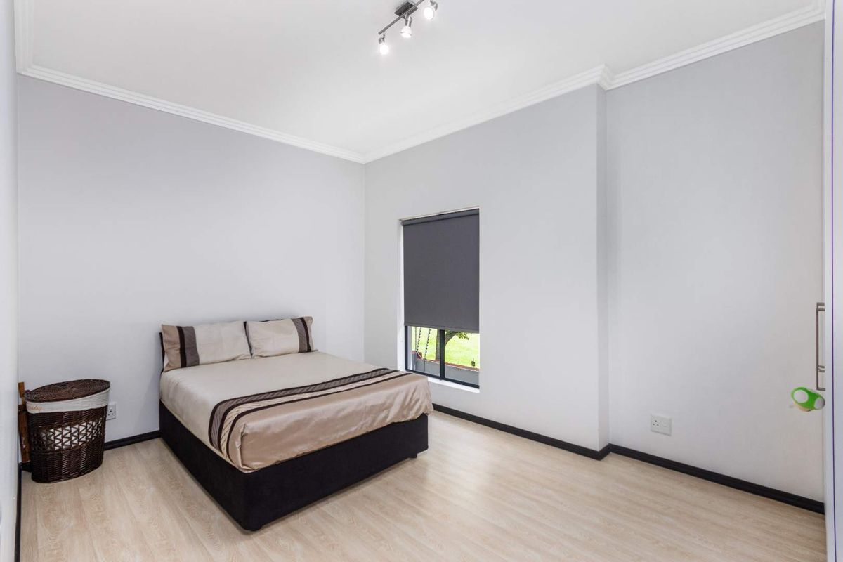 A bright and neutral bedroom featuring light laminate flooring, clean white walls, ceiling cornice detailing, and a modern roller blind.