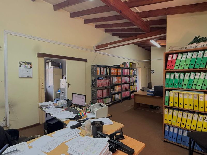 A functional office space featuring exposed timber ceiling beams and carpeted flooring, currently utilized as a workspace with storage shelving.