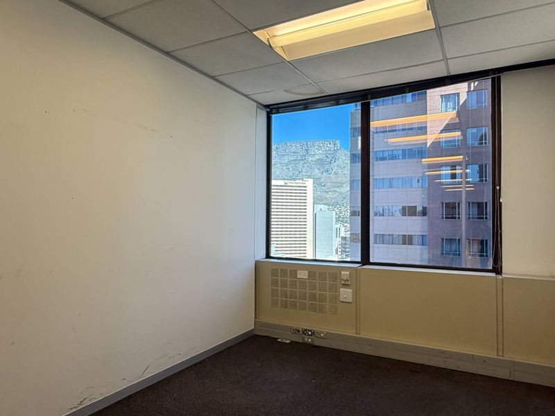 An empty office space featuring a suspended ceiling, carpeted flooring, and a large window offering city and mountain views.