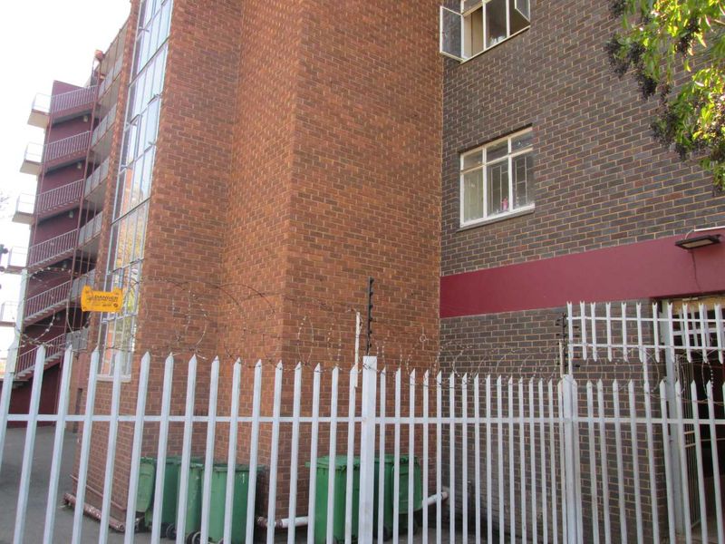 Exterior view of the apartment building featuring a brick facade, steel fire escape staircase, and secure perimeter palisade fencing with electric wires.