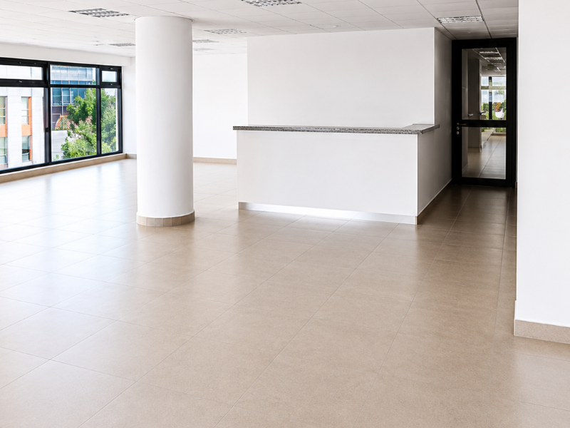 A bright, open-plan office space featuring neutral ceramic floor tiles, a central structural column, a built-in reception counter, and large windows providing natural light.