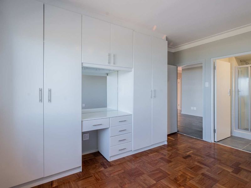 A bright bedroom featuring classic parquet flooring, extensive built-in white cabinetry with an integrated vanity, and direct access to an en-suite bathroom.