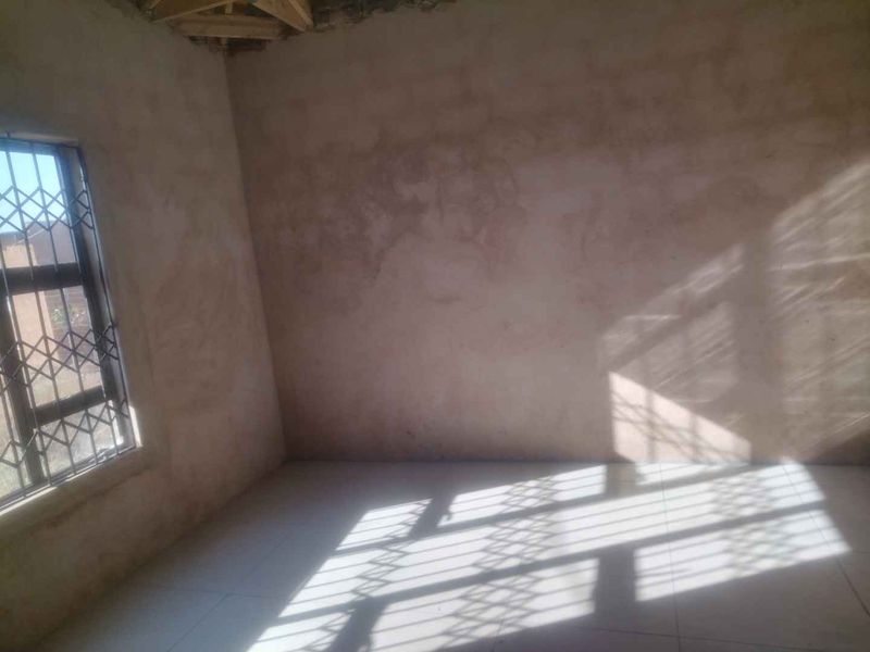 An unfinished bedroom space featuring tiled flooring, a security gate on the window, and exposed roof trusses, requiring final plastering and ceiling installation.