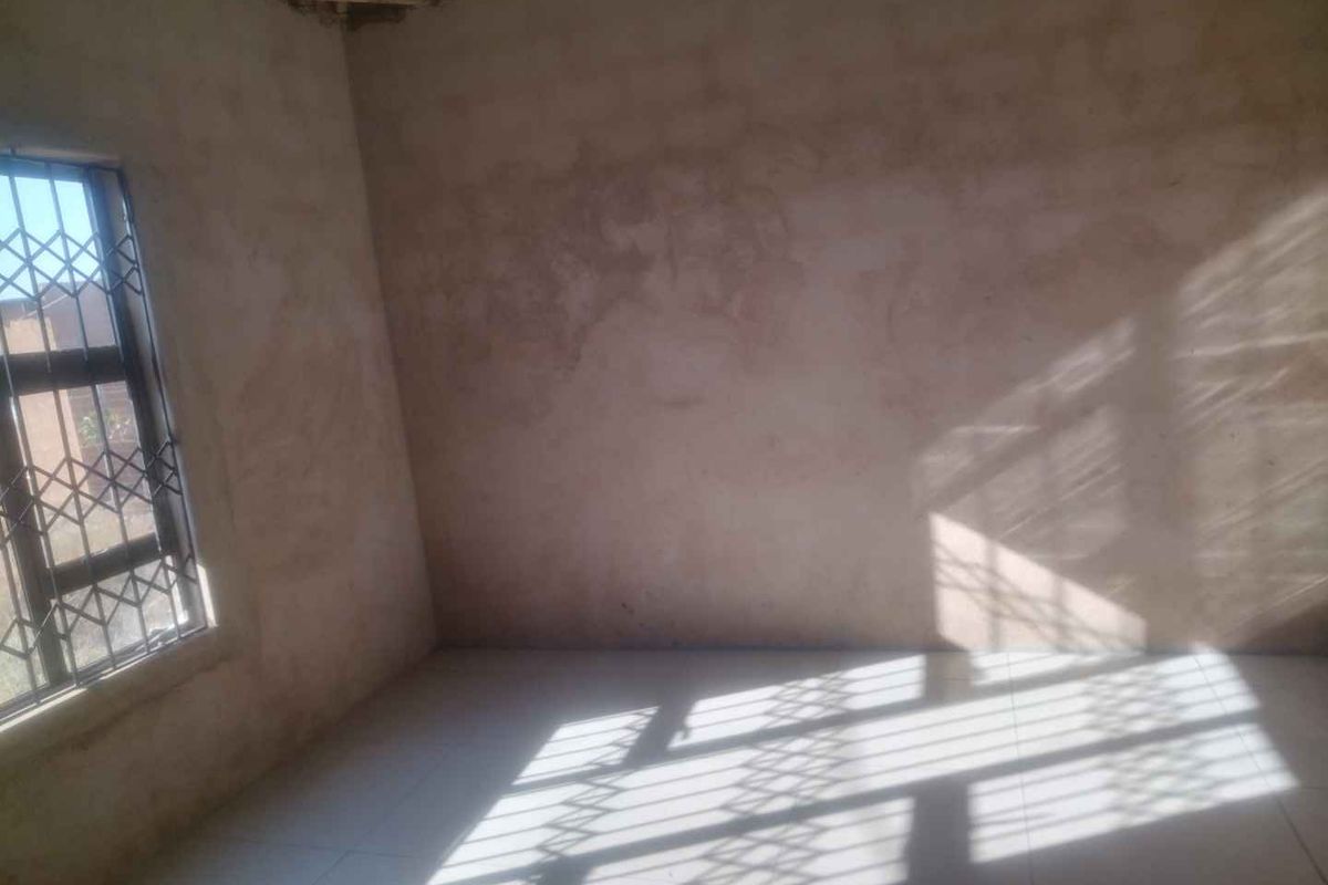 An unfinished bedroom space featuring tiled flooring, a security gate on the window, and exposed roof trusses, requiring final plastering and ceiling installation.