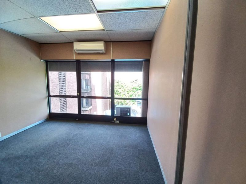 A vacant office space featuring a drop ceiling, wall-mounted air conditioning unit, and large windows providing natural light.
