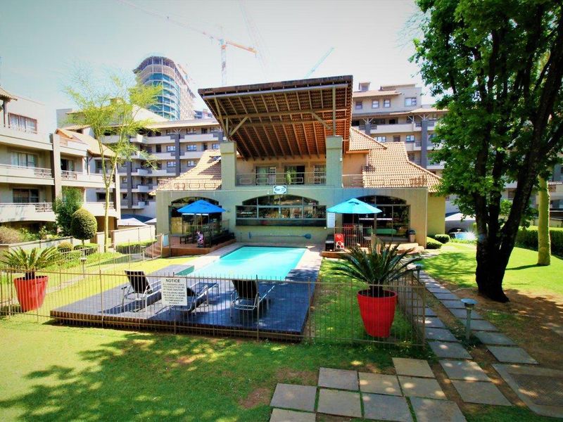 A well-maintained communal swimming pool area featuring a paved deck, metal fencing, and landscaped gardens, situated within an apartment complex.