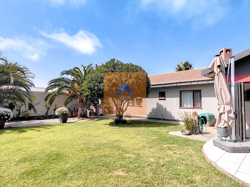 A well-maintained rear garden featuring a lawn, mature palm trees, and a tiled patio area attached to the main house structure.