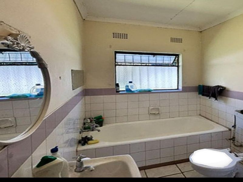 A dated bathroom featuring a built-in bathtub, pedestal basin, and toilet, with tiled splashbacks and a window for natural light.